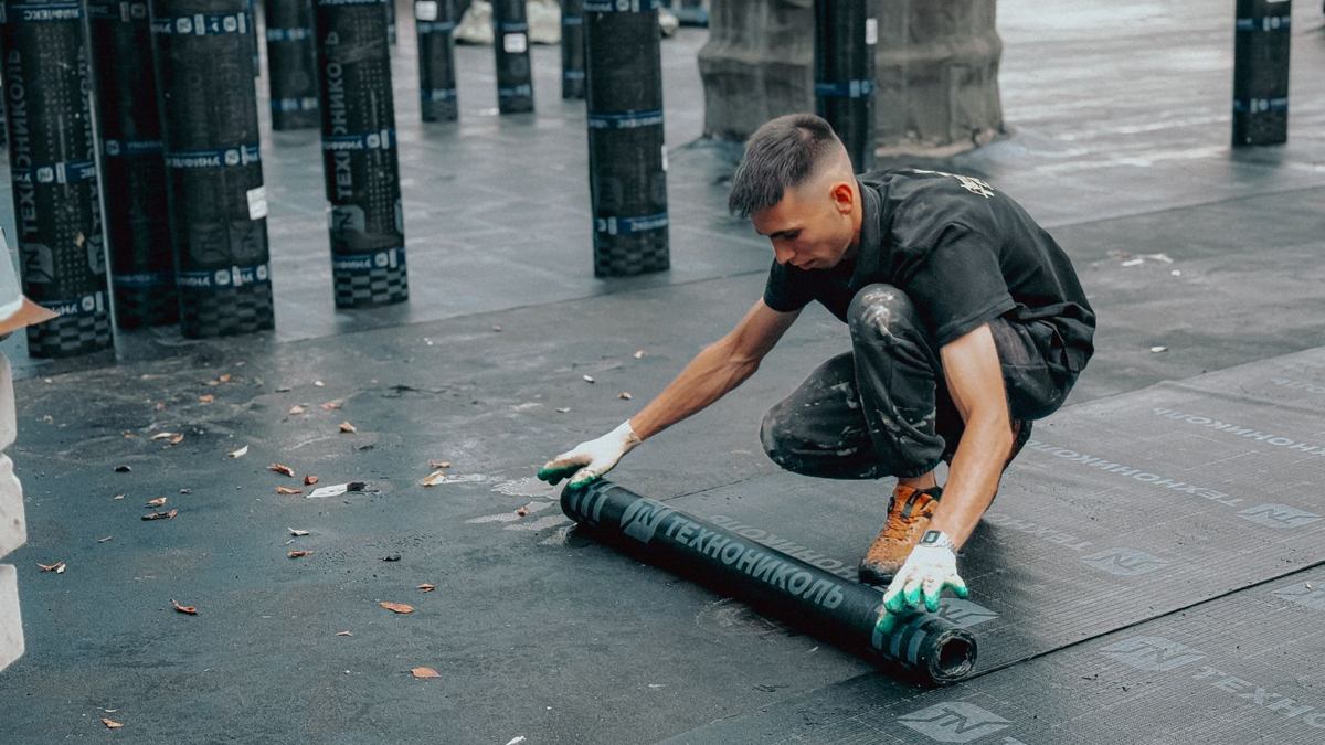 Waterproofing contractor in gloves kneeling to roll out a heavy black membrane across a flat surface
